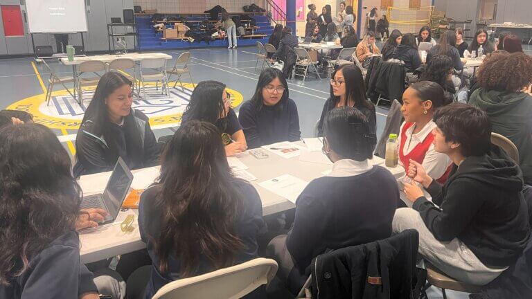 Volunteers and students are seated around long tables, with laptops and papers in front of them.