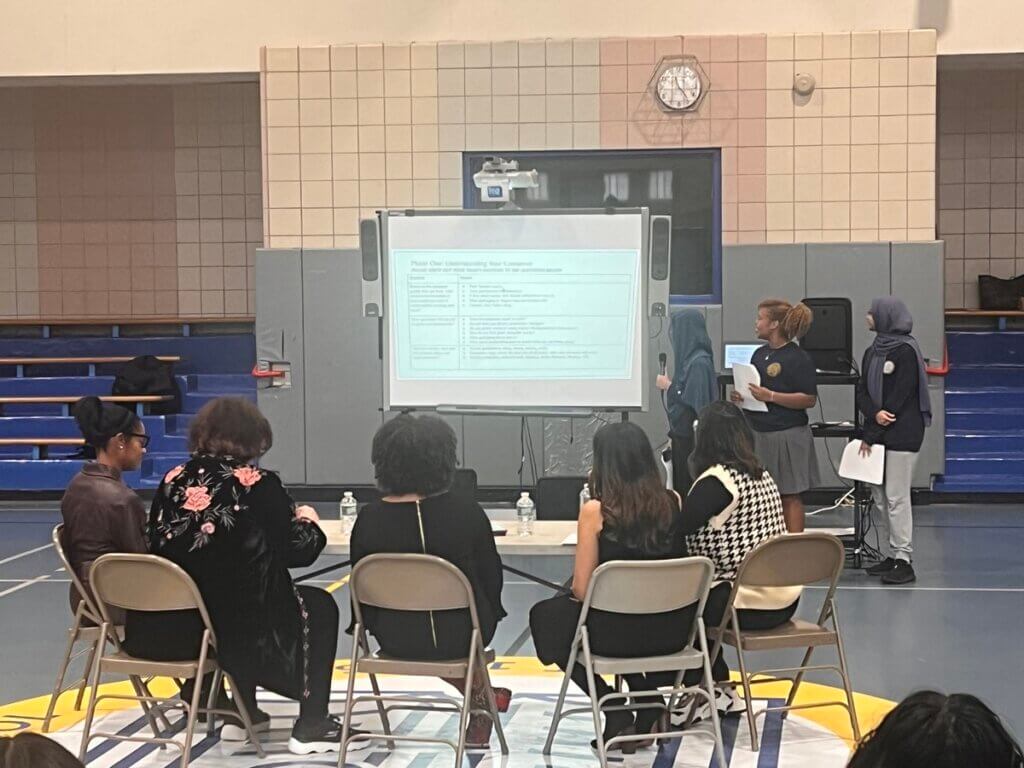 Three students explain a chart on a screen to a panel of pitch competition judges