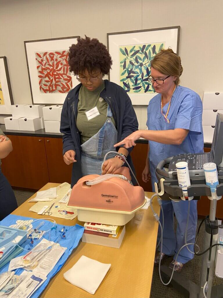 Student holds a sensor attached to a sonogram machine while a medical faculty member next to her points to the model dummy