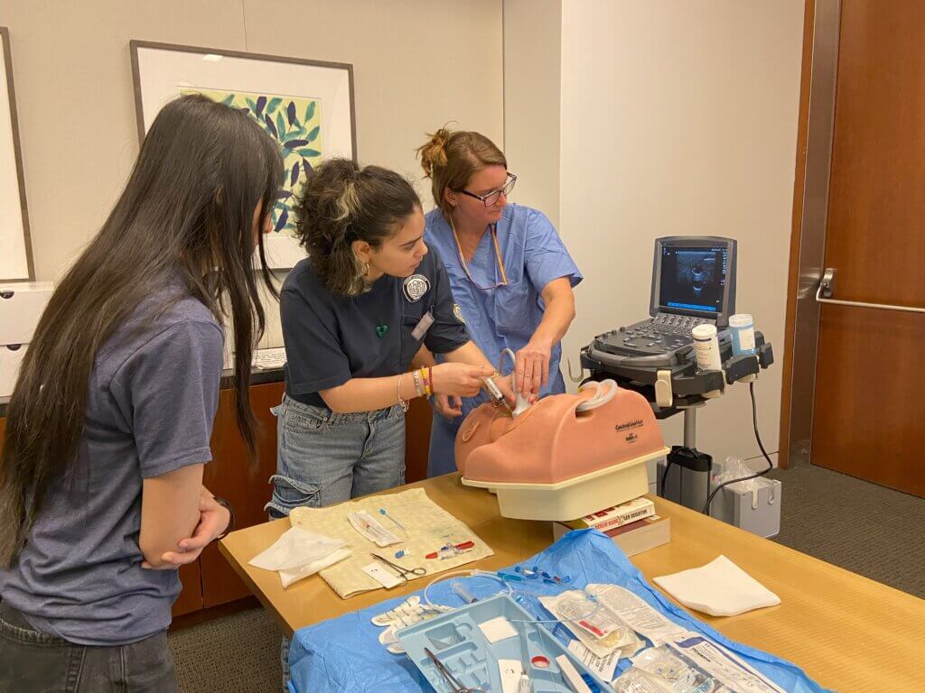 Student holds a needle while a medical faculty member guides the sensor attached to a sonogram machine