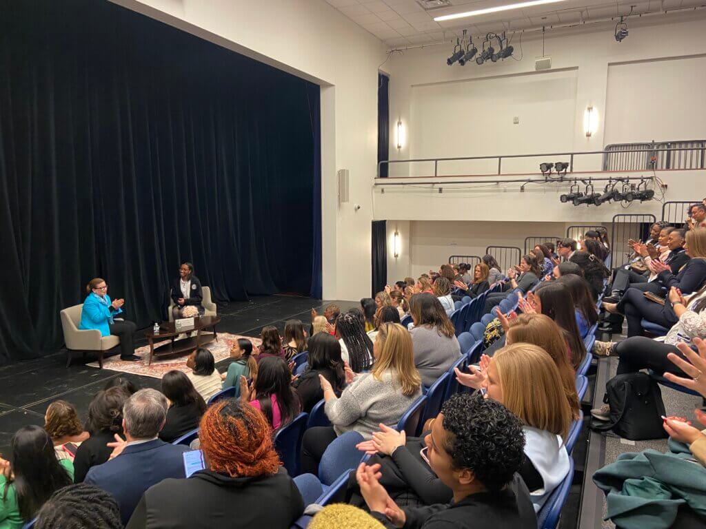 Valerie Jarrett and student Sariyah seated on stage, with audience clapping