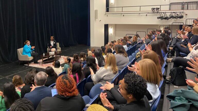 Valerie Jarrett and student Sariyah seated on stage, with audience clapping