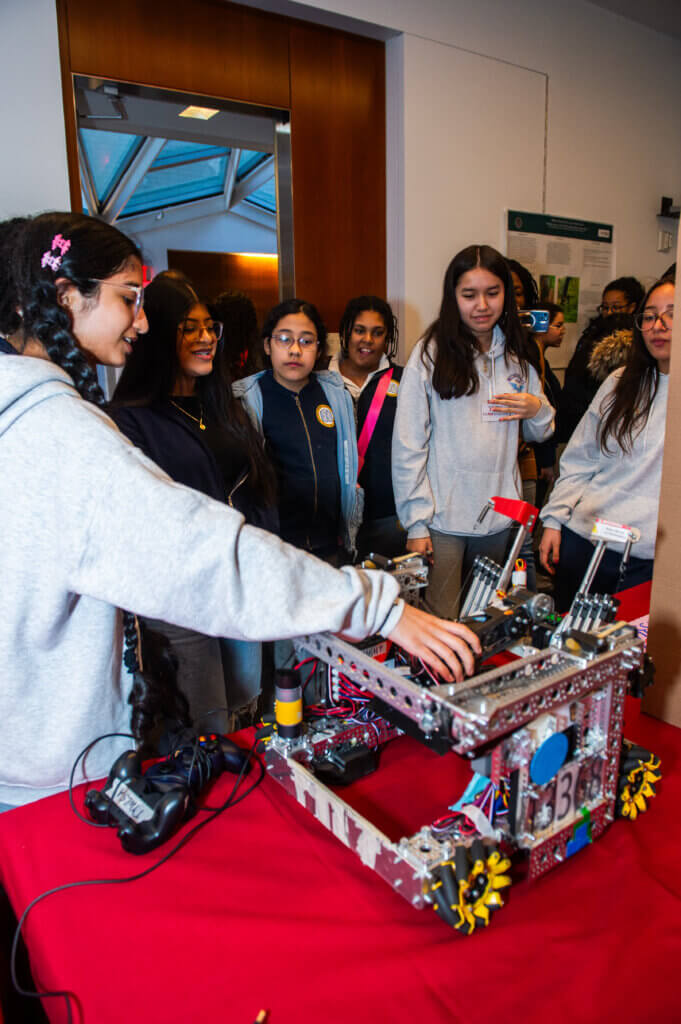 Student points to different parts of a robotics machine while a group of students gather around her