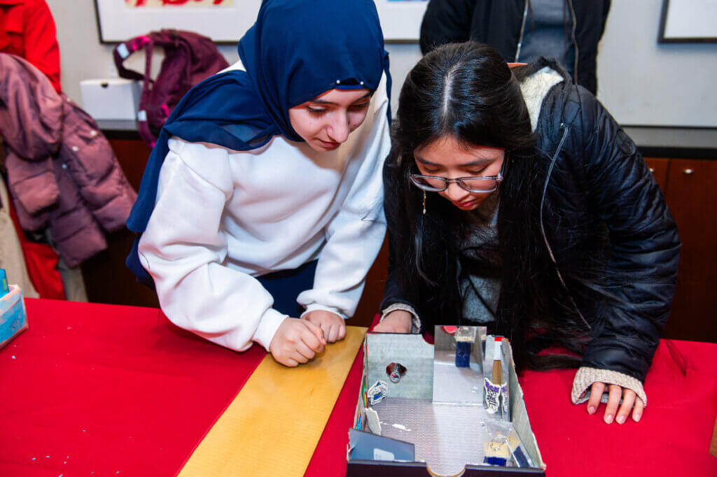 Two students lean in to examine a reflective cardboard box on the table