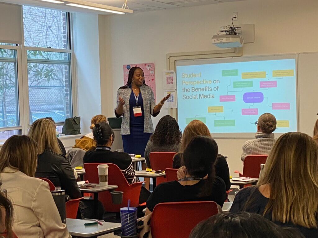Woman standing gestures to a screen in front of classroom, while seated adults look on