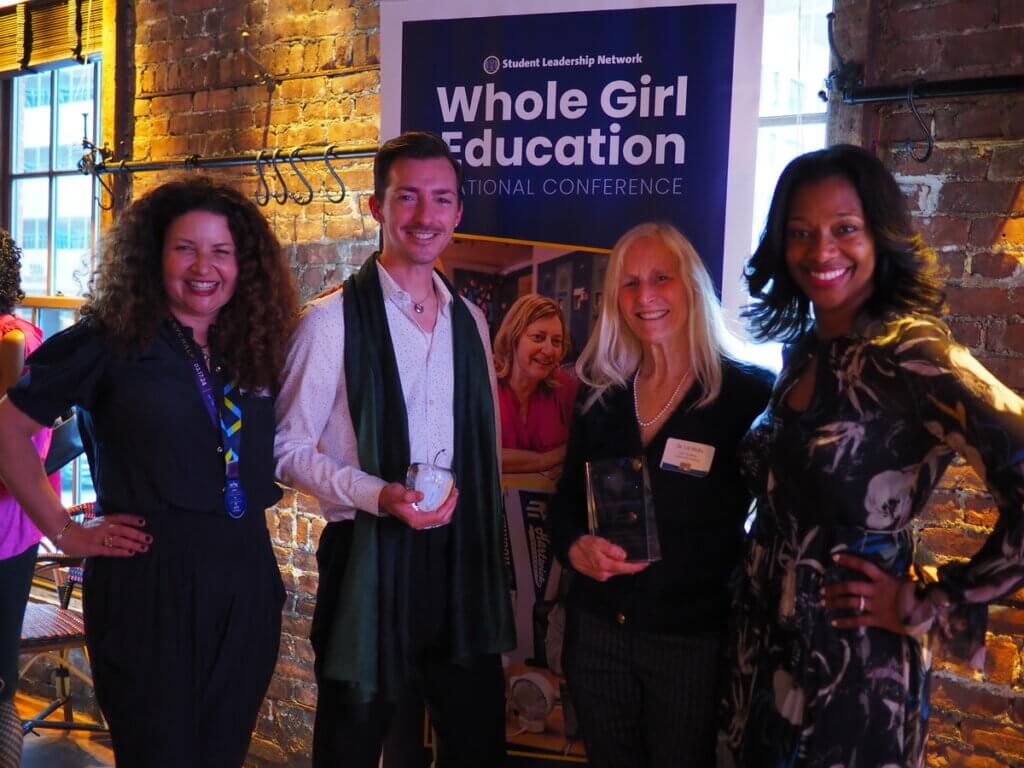 Four adults pose in front of a banner, with two educators in the center holding up crystal awards