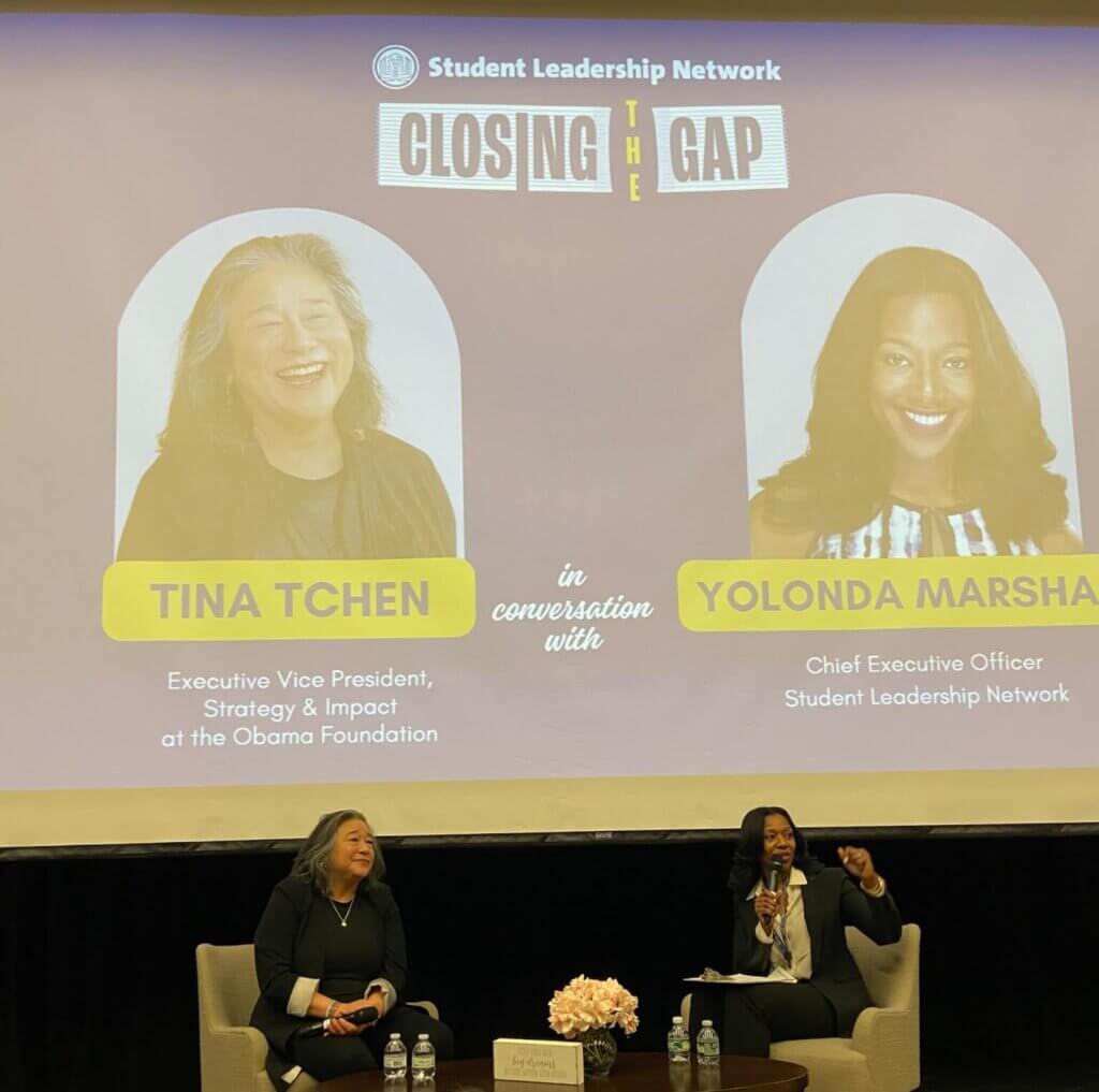 Two women are seated on a stage with microphones, speaking to audience