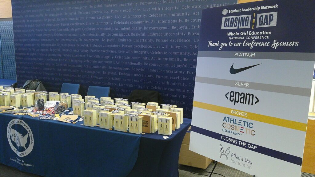 Poster board on right with sponsorship logos of Nike, EPAM, Athletic Cosmetic Company, and Riley's Way; next to table of in-kind donations and giveaways