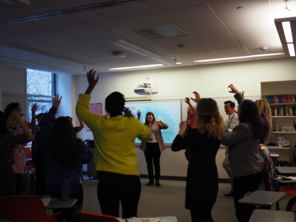 Adults stand in circle for an improv activity, with arms raised in the air