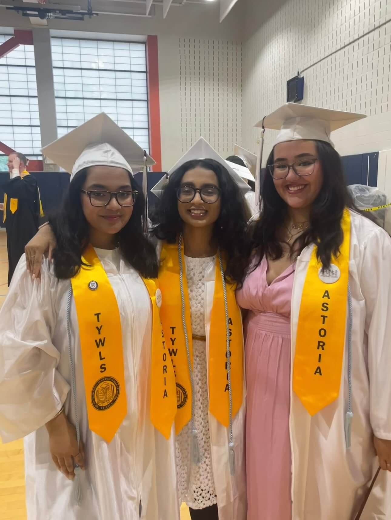 Three young women wearing graduation caps and gowns with TYWLS Astoria stoles across their shoulders