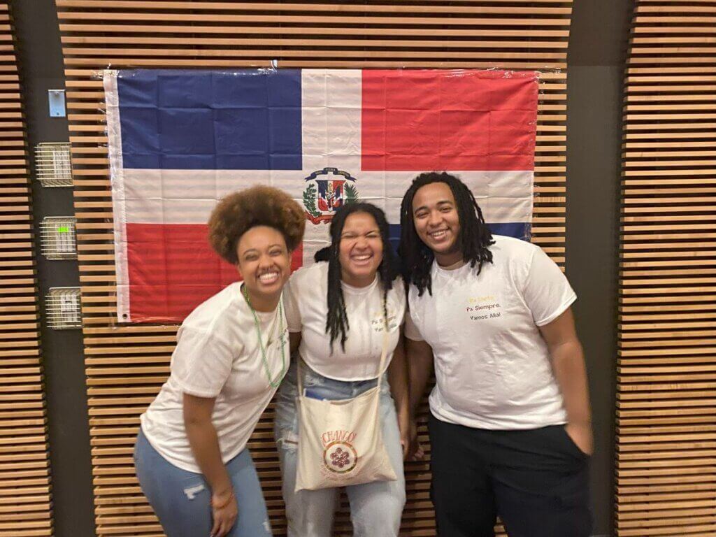 Three college students smile in front of a flag of the Dominican Republic