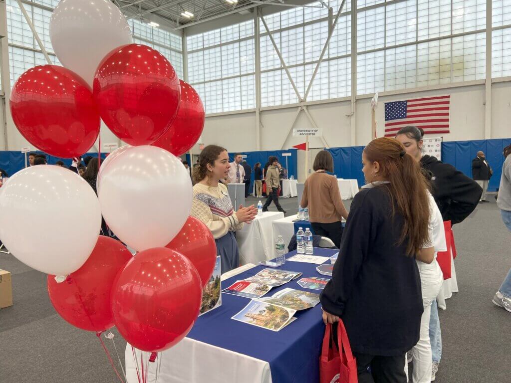High school students visit a variety of college admissions tables inside a large gymnasium