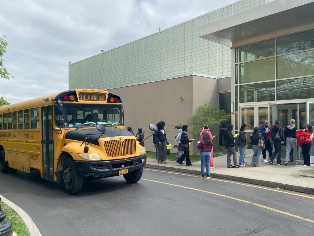 Yellow school bus in front of glass building, as students disembark and walk inside