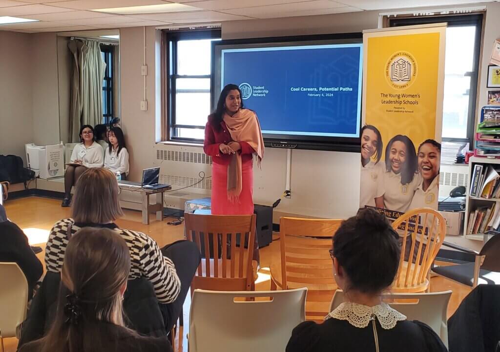 Principal stands in front of room to welcome volunteers, adults are seated in front of her and students on the side
