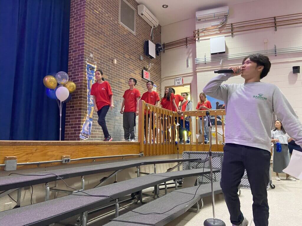 student talks into microphone as other students in red shirts walk across stage
