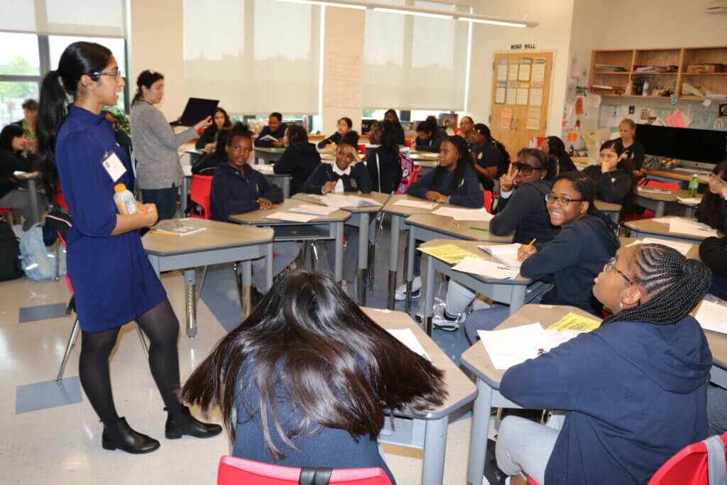 Adult volunteers stand on left side of classroom while students are seated in semi-circles