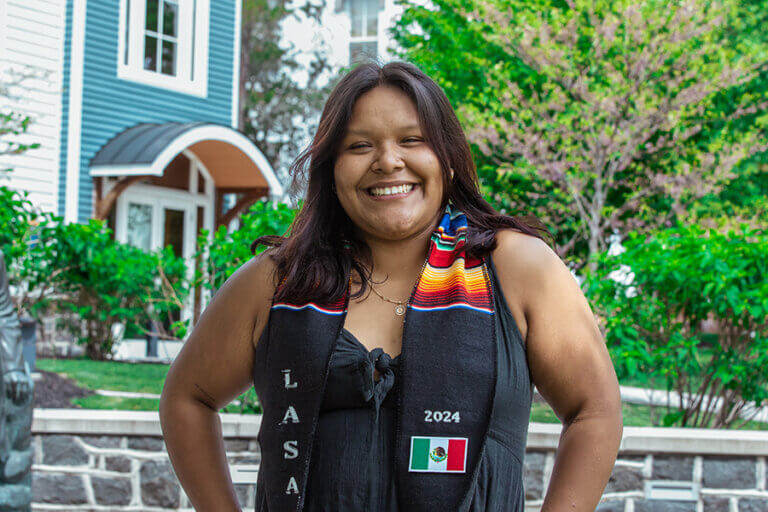 Young woman wearing a graduation stole with "LASA 2024" and the Mexican flag on it stands in front of a tree-lined street