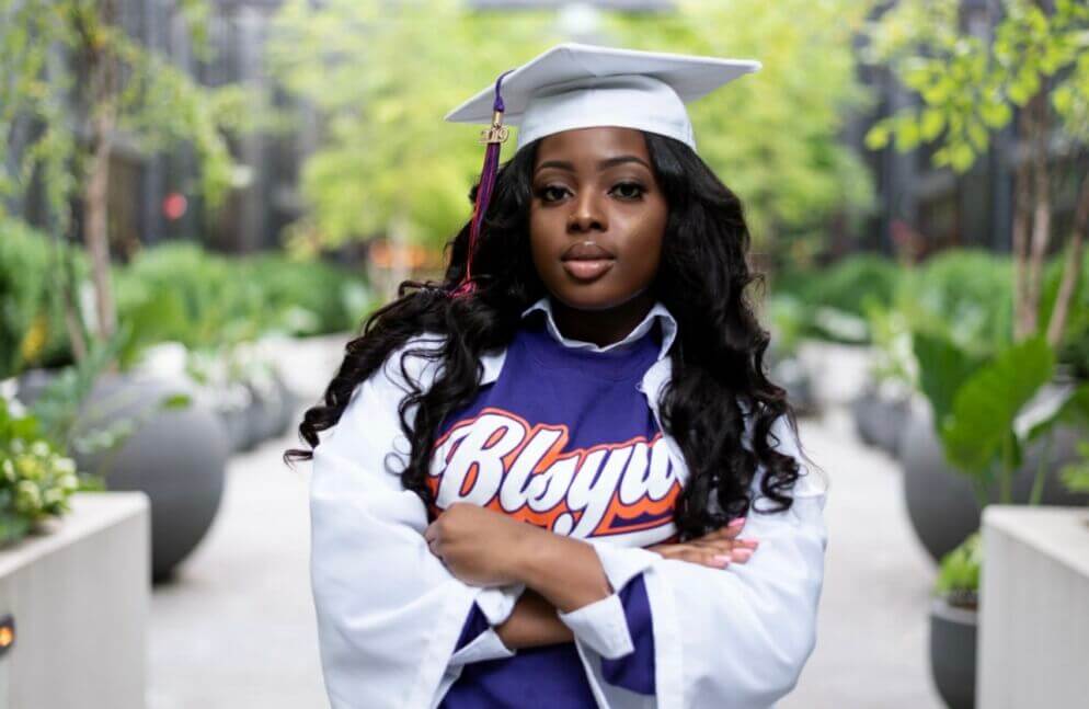 Young woman wears a white graduation cap and gown, with purple t-shirt with text "BLSYW" across the front