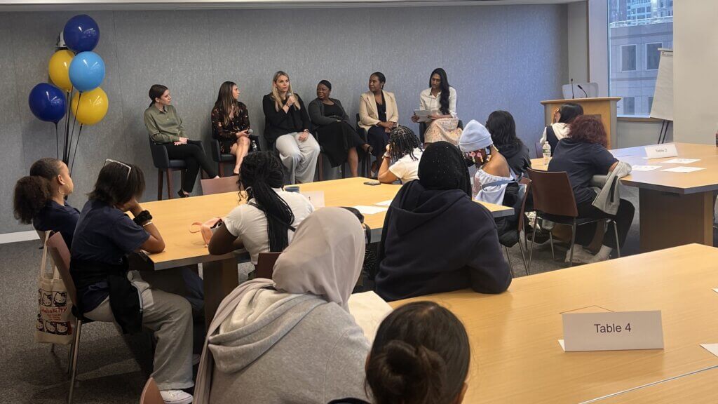 Women panelists are seated at front of room with microphones, while students are seated at conference tables