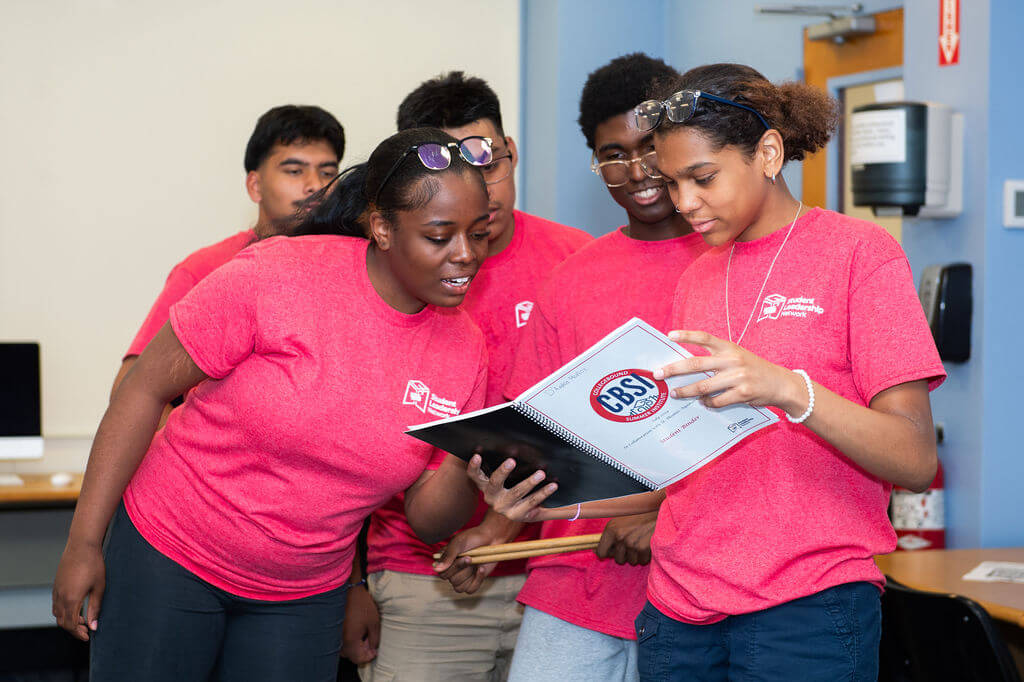 Students in red shirts crowd around a spiralbound notebook that says "CBSI" on the cover