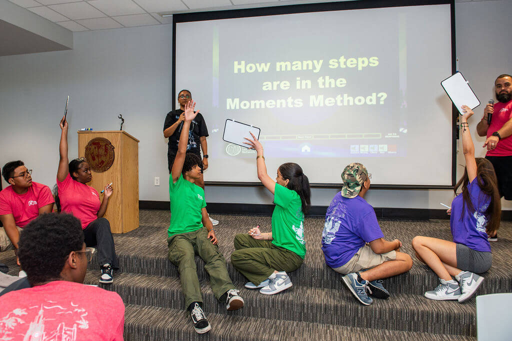 Six students sit on stage, holding up dry-erase boards, while two counselors ask Jeopardy-style questions