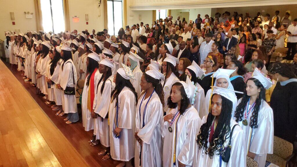 High school students in graduation caps and gown stand in auditorium, with parents, family, and friends behind them