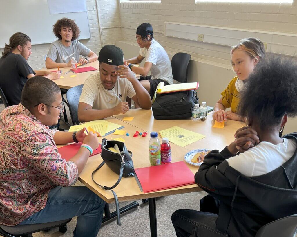 Four students are seated in around table, with folders and essay papers in front of them