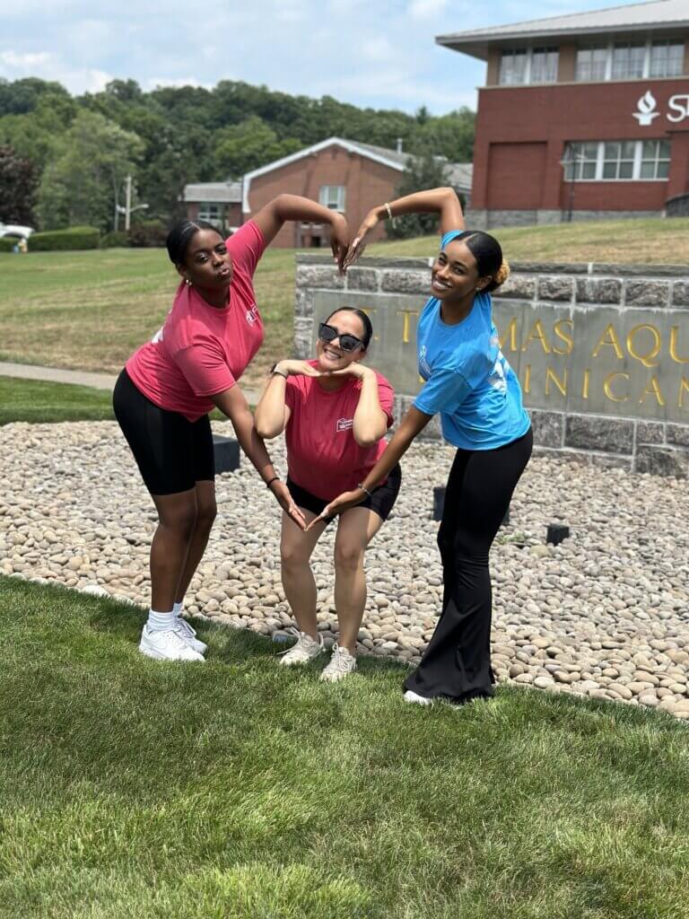 A counselor and two of her students pose for a photo outdoors, their arms making a heart shape