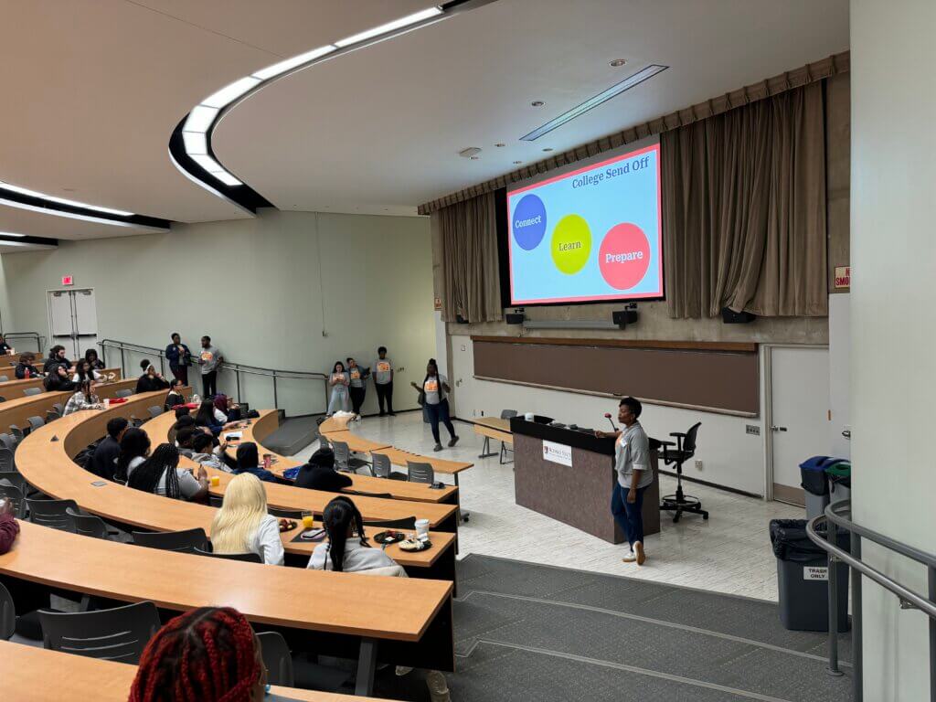 Students sit in half-circles in a college lecture hall, with text "College Send Off" projected on the screen
