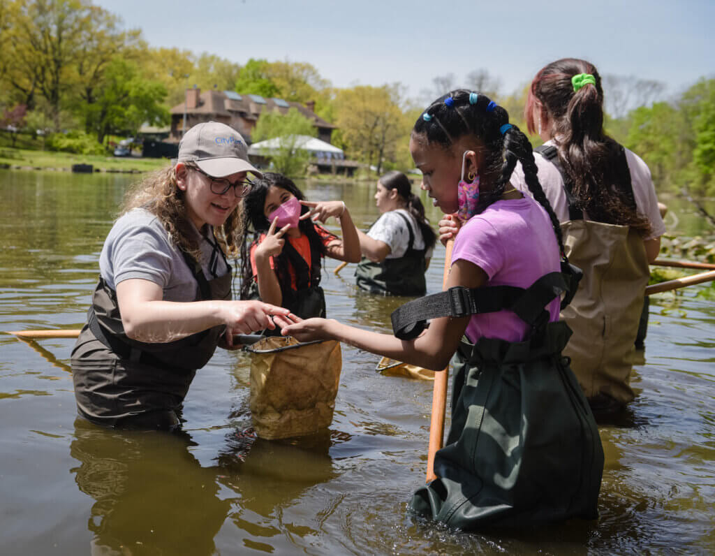Four girls wearing waterproof coveralls are in a pond gathering plants and insects