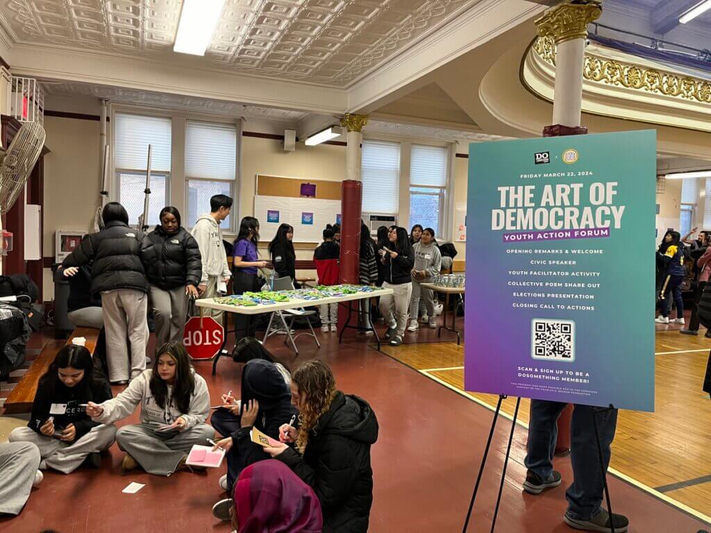Groups of girls sit in circle, with table of post-it notes, with Art of Democracy poster board in foreground