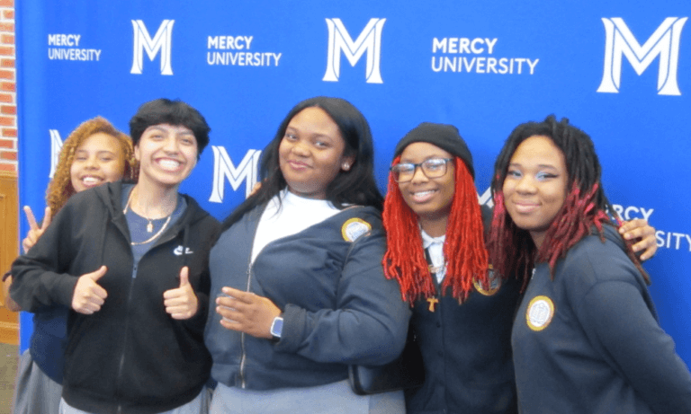 Five students stand in front of blue backdrop with Mercy University signage