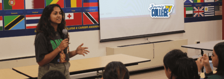 A young woman shares college application tips for parents, speaking into a microphone in front of an audience in a classroom decorated with international flags and a Journey to College sign by the projector screen.
