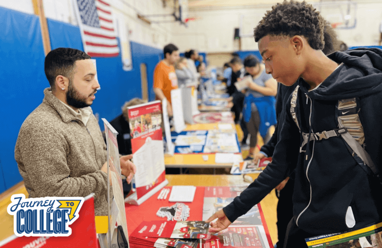 Young man with backpack reaches for a college brochure while college rep speaks from behind table