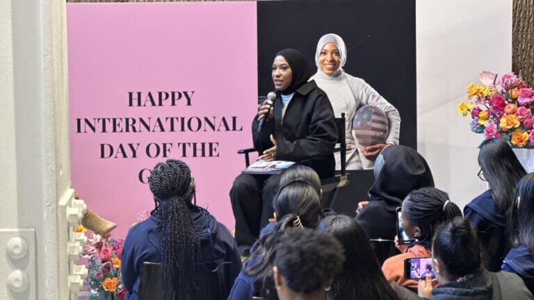 A woman in a hijab speaks into a microphone at an event celebrating International Day of the Girl, with an audience seated in front of her and a floral display beside the stage.