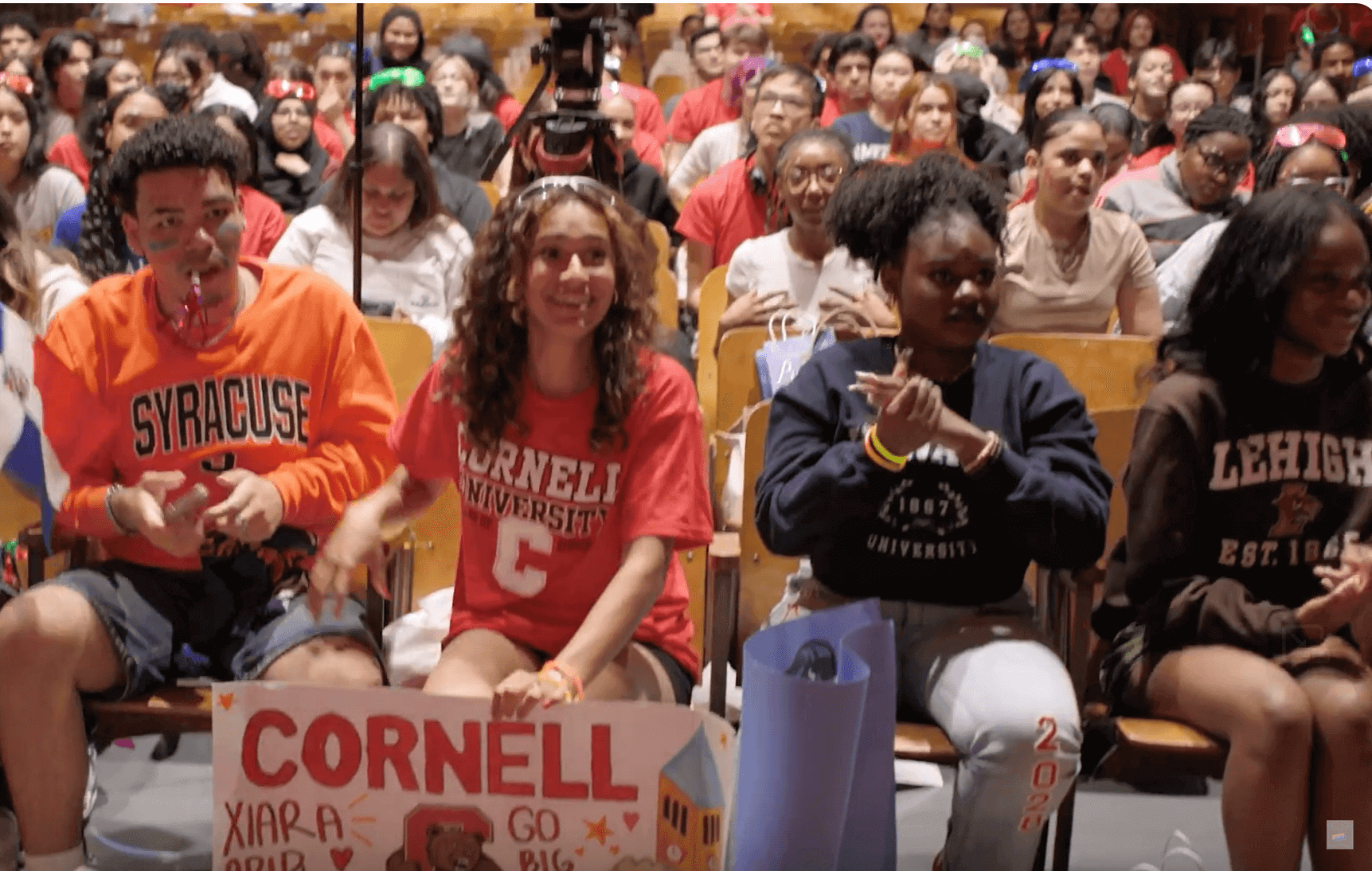 At the 2024 EmPower Breakfast Showcase, a group of students sits in an auditorium; three in front wear Syracuse, Cornell, and Lehigh shirts, and one holds a Cornell sign. The audience watches attentively as College Matters is celebrated.