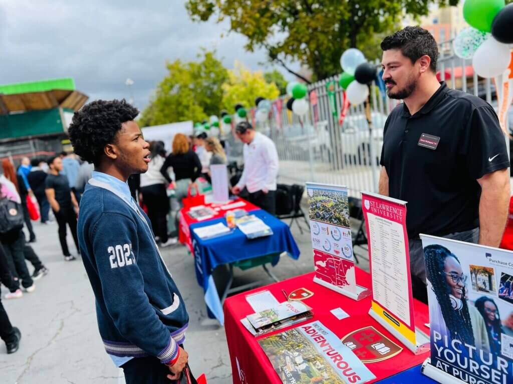 Student wearing a knit cardigan talks with a college admissions rep by a table of college brochures
