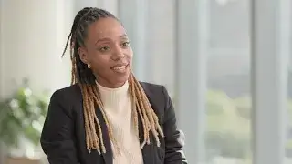 A woman with long, light brown braids—2024 Honoree Kenya Bryant—wears a black blazer and light-colored top as she smiles indoors at a Power Breakfast, with large windows and greenery in the background.