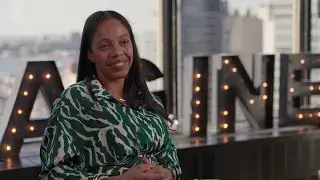 A woman with long dark hair wearing a green and white patterned blouse sits indoors, smiling, with large lit-up letters spelling IMAGINE in the background at the Power Breakfast honoring Sharlim Raysor as a 2024 Honoree.