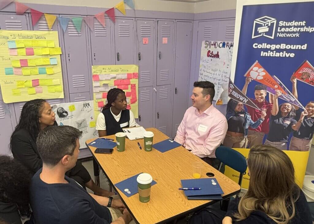Adult volunteers and students are seated and talking inside a classroom
