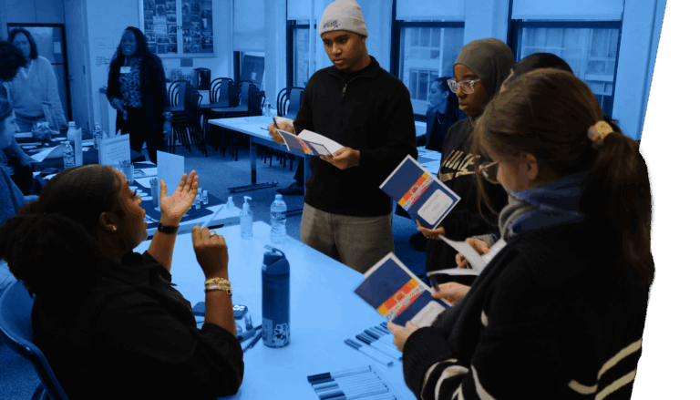 four students take notes on paper as a woman seated at an event table speaks with them