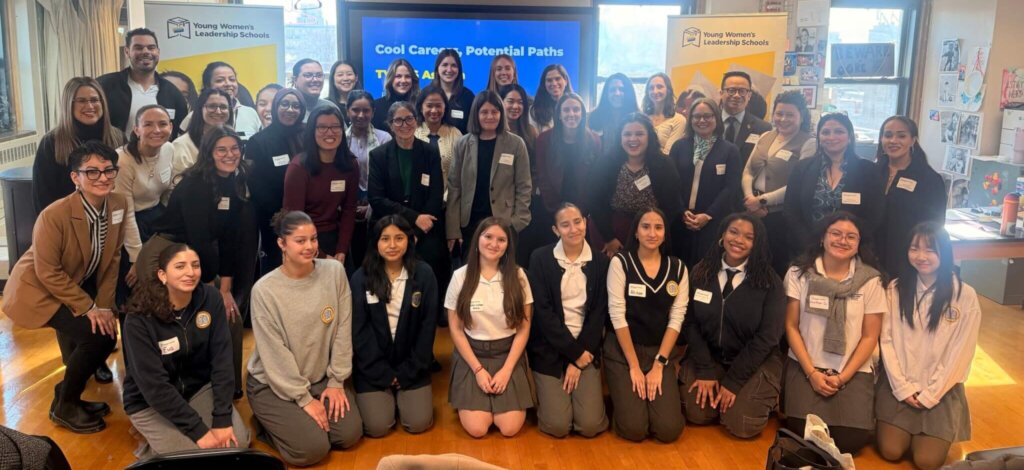 A large group of young women and adults pose together indoors in front of a presentation screen and banners, dressed in business and school attire, gathered to celebrate volunteer appreciation week.