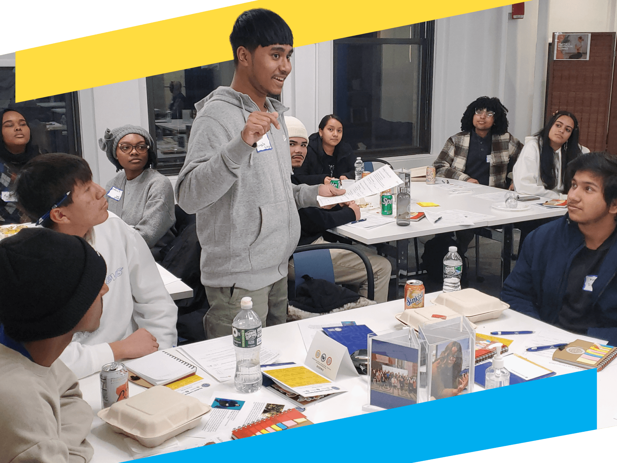 A young man, part of the Student Leadership Network alumni services, stands and speaks in a classroom setting while others sit around tables listening attentively