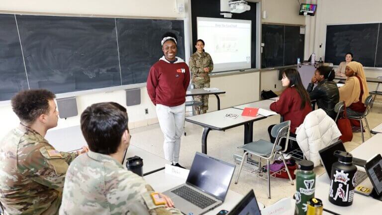 Female student stands smiling, facing the classroom while students and military personnel are seated in front of her