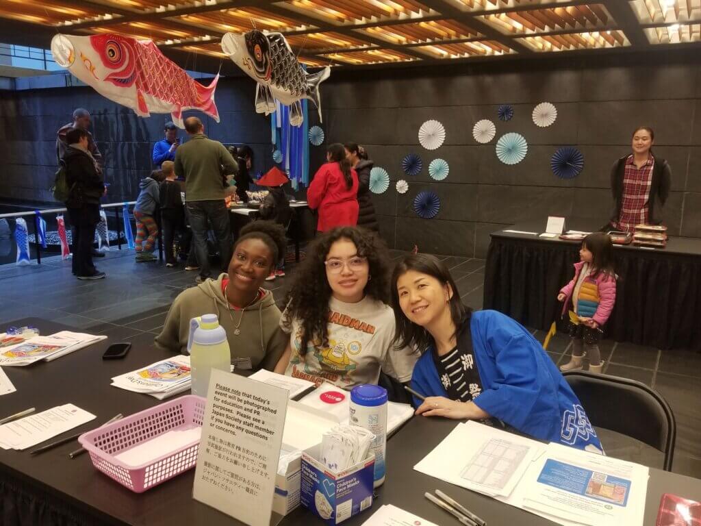 Two students and an adult are seated behind a check in table of a museum, with fish-shaped paper kites hanging from the ceiling