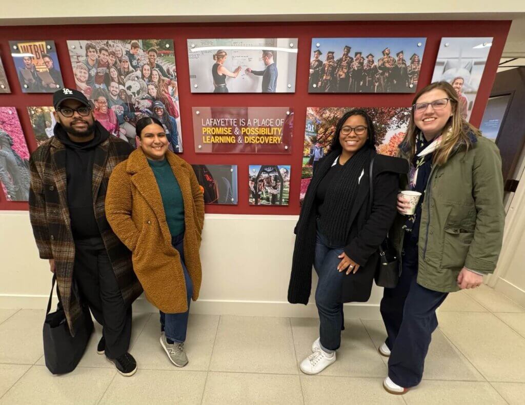 Four people stand indoors in front of a bulletin board displaying photos and a sign that reads Lafayette is a place of promise & possibility, learning & discovery, celebrating the SUNY McConney EOP award.