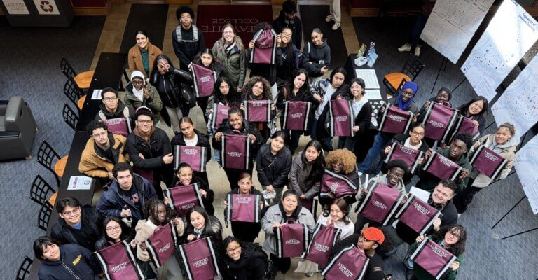 A large group of students stands indoors in a circle, holding maroon tote bags and looking up at the camera, celebrating their SUNY McConney EOP award. Tables and chairs are visible in the background.