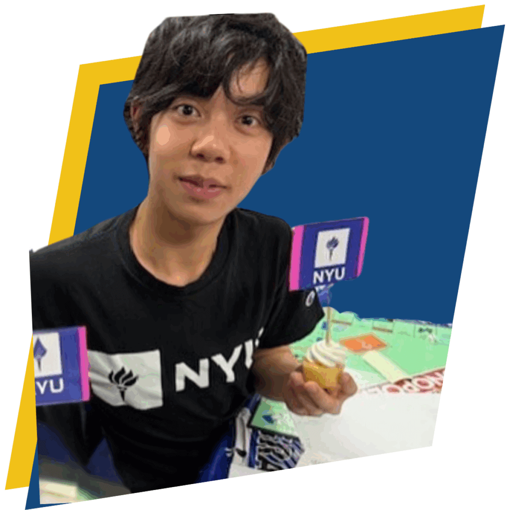 Young person wearing an NYU shirt, symbolizing student leadership, holds a small cupcake with an NYU flag on it while sitting at a table with papers and another NYU flag visible.