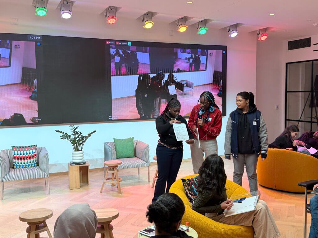 Three people stand at the front of a modern room presenting a paper to an audience, with a large screen behind them displaying their image.
