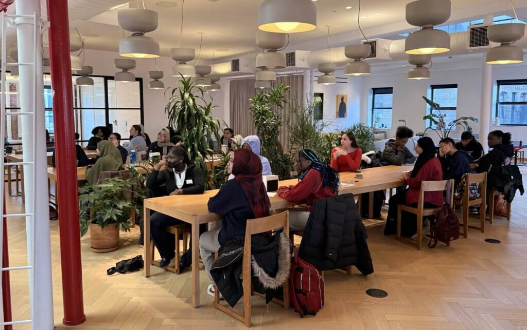 A group of people sit at long tables in a modern, brightly lit room with hanging lights and indoor plants, attending what appears to be a meeting or workshop.
